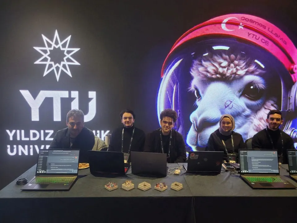 Five people seated at a table with laptops in front of a large Yıldız Technical University display showing a llama astronaut