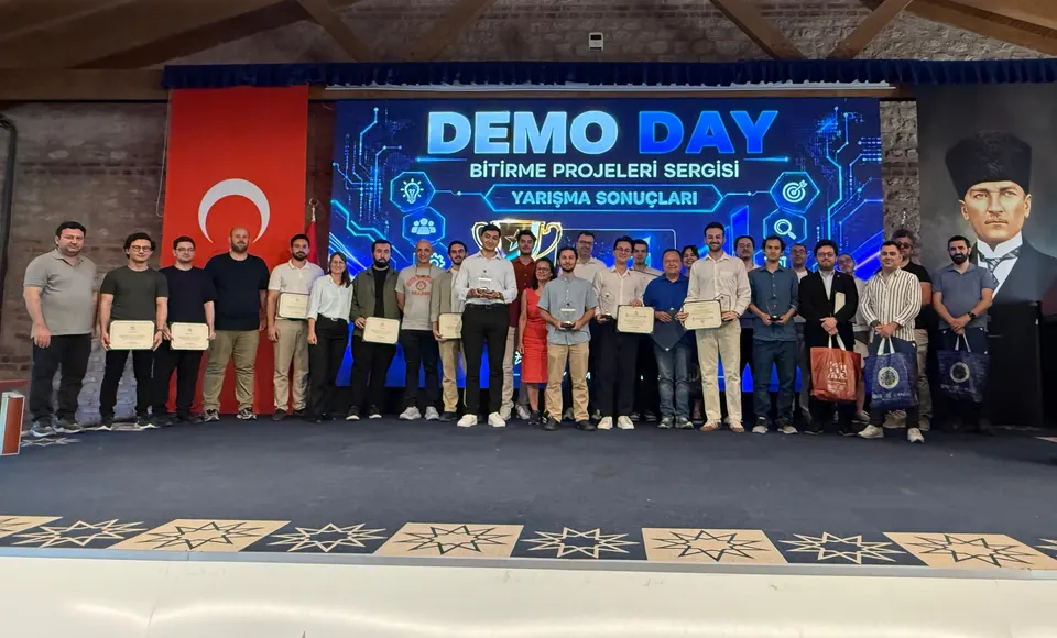 Large group posing with certificates on stage under a "Demo Day" screen at a graduation projects exhibition