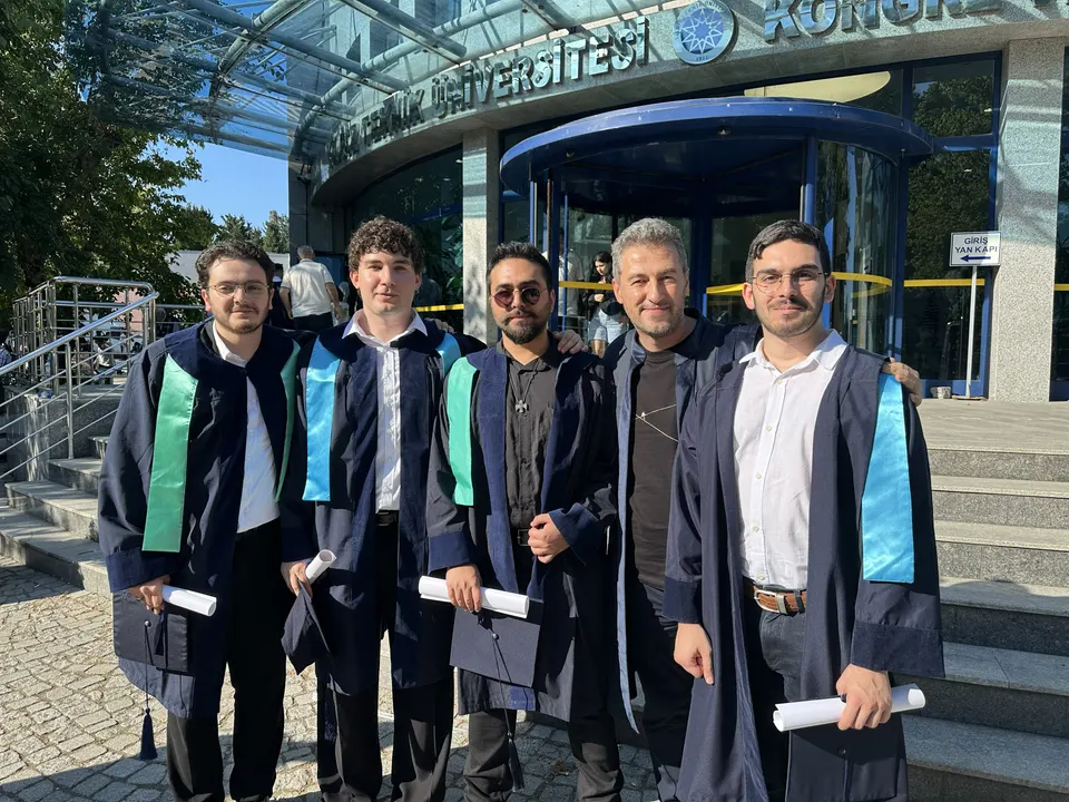 Group of five in graduation gowns, most holding diploma scrolls, in front of a university building